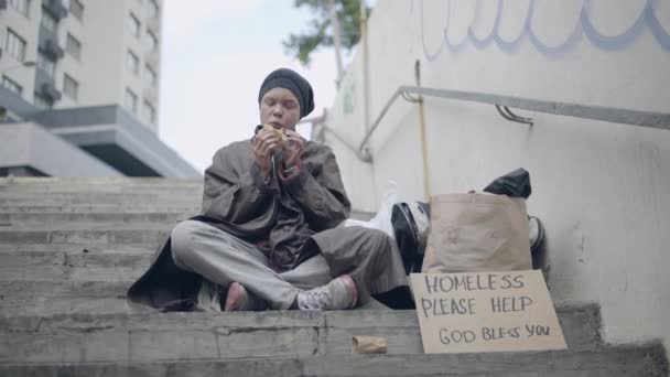 Homeless young female eating sandwich, sitting on the stairs, begging for food — Stock Video