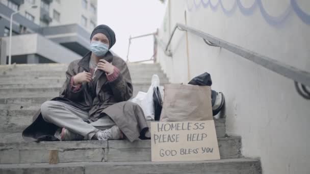 Одинокая бездомная женщина в маске, глядя в камеру, помогите расписаться на лестнице — стоковое видео