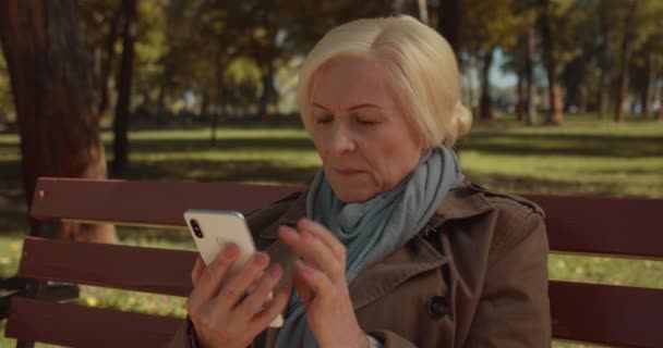 Bezorgd senior vrouwelijke lezing schokkend nieuws op smartphone, zitten in het park — Stockvideo