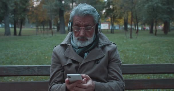 Homem moderno em fones de ouvido ouvindo música no parque, usando o aplicativo do smartphone — Vídeo de Stock