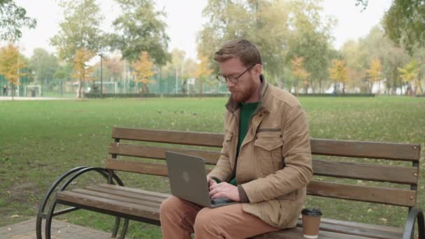 Мужчина-блогер в очках печатает на ноутбуке, сидит в парке с чашкой кофе — стоковое видео