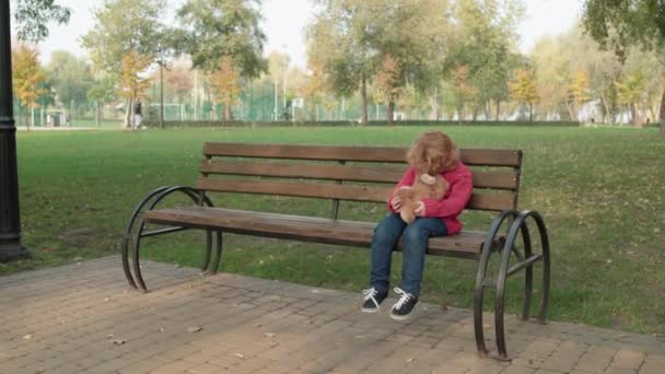Потерянный рыжий мальчик сидит на скамейке в парке, держит медвежью игрушку, чувствует себя одиноким — стоковое видео