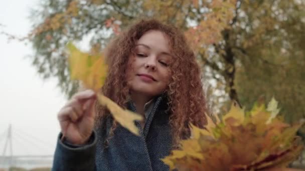 Young pretty female with curly red hair holding autumn leaves, relax in forest — Stock Video