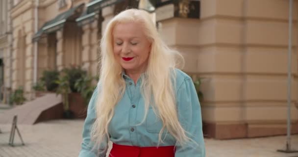 Радісна довгошерста зріла жінка в стильному вбранні перевіряє сумки на відкритому повітрі — стокове відео