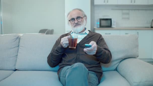 У віці чоловік дивиться телевізор вдома, п'є чай на дивані, розслабляється у вільний час, хобі — стокове відео