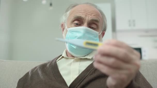 Грустный пожилой человек в защитной маске показывает термометр, симптомы лихорадки, вирус — стоковое видео