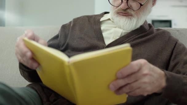 Pensionista relajante en casa, sentado con libro en el sofá, tiempo libre, conocimiento — Vídeo de stock