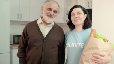 Alışveriş çantasıyla mutlu kadın gönüllü ve kameraya gülümseyen yaşlı adam, yardım et.
