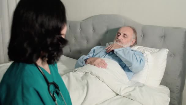 Молода медсестра дивиться на термометр, піклуючись про літнього пацієнта вдома — стокове відео