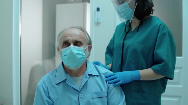 Enfermeira em máscara e protetor facial examinando os pulmões do paciente, usando estetoscópio — Vídeo de Stock