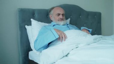 Yaşlı bir adam hastane yatağında battaniyenin altında yatıyor, huzurevinde yaşlılara bakıyor.