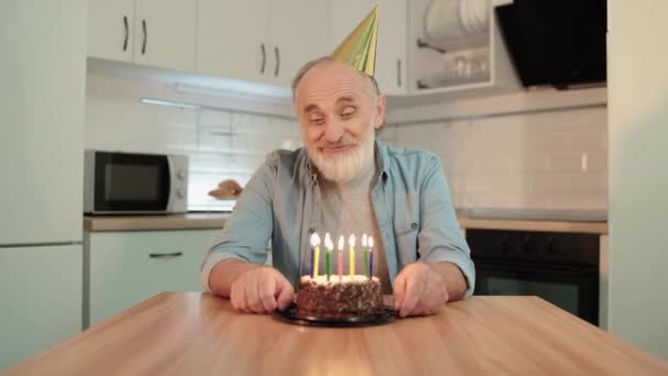 Uśmiechnięty starzec w imprezowym stożku wypowiadający życzenie, zdmuchujący świeczki, urodziny — Wideo stockowe