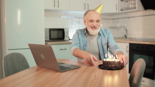 Szczęśliwy stary człowiek w imprezowym kapeluszu zdmuchnąć świece, rozmawiając z krewnymi online — Wideo stockowe