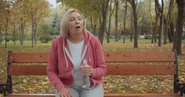 Trött senior kvinna dricksvatten från flaskan efter jogging i parken, förfriskning — Stockvideo