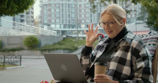 Mujer sonriente en anteojos y auriculares haciendo videocall portátil, saludando de la mano — Vídeos de Stock