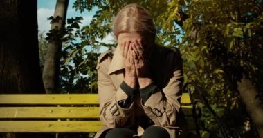 Üzgün yaşlı kadın parktaki bankta tek başına ağlıyor, acıdan ellerini sıkıyor.