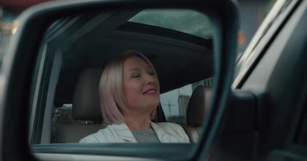 Улыбающаяся блондинка 50-ти лет, регулирует прическу, смотрит в зеркало автомобиля — стоковое видео
