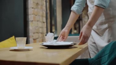Kadın bistro işçisi kirli tabak ve bardakları alıyor, masayı temizliyor.