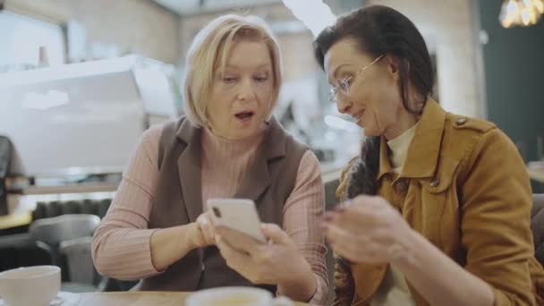 Współcześni starsi przyjaciele oglądają zdjęcia na smartfonie, siedzą razem w kawiarni — Wideo stockowe