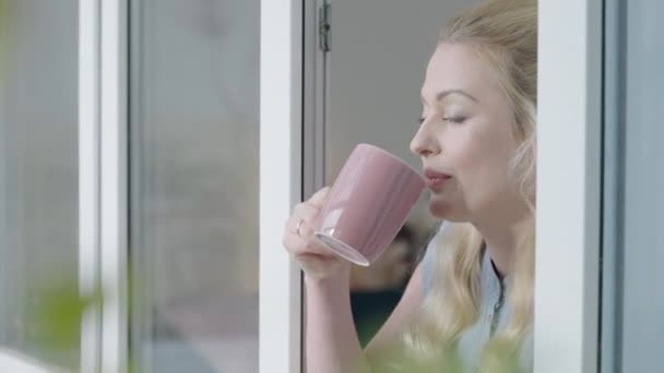 Vidám szőke nő kávézik reggel, az ablak mellett áll, betör — Stock videók