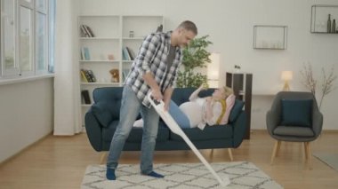 Koca yerleri süpürüyor, hamile kadın kanepede dinleniyor, yeşil elma yiyor.