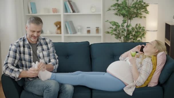 Schwangere entspannt sich auf Sofa, telefoniert, liebender Ehemann massiert Füße — Stockvideo