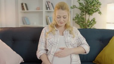 Hamile bir kadının karnını okşaması, kameraya gülümsemesi, evdeki kanepede oturması.