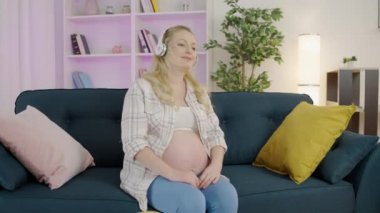 Sarışın, hamile karnına kulaklık takan, bebekle bağ kuran bir kadın bekliyor.