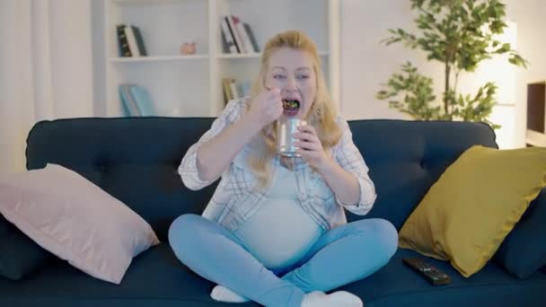 Fiatal terhes nő eszik konzerv kukorica, TV-t néz kanapén, terhesség utáni sóvárgás — Stock videók