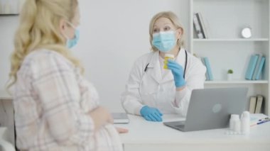 Maskeli jinekolog hastaya doğum öncesi bakımı sırasında doğum kontrol haplarını gösteriyor.