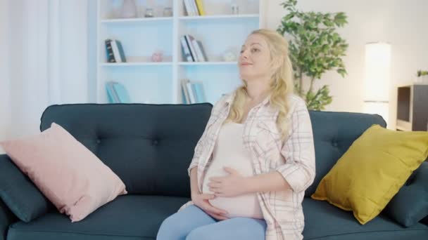 Улыбающаяся беременная женщина гладит живот, сидит на диване, чувствует себя счастливой, гармоничной — стоковое видео