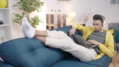 Kulaklık takan, dizüstü bilgisayarda yazan, bir proje üzerinde çalışan, kanepede dinlenen genç bir adam.