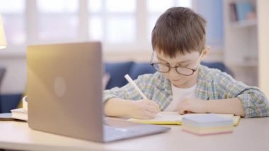 Gözlüklü öğrenci ödevlerini yapıyor dizüstü bilgisayarın önünde oturuyor, uzaktan öğreniyor.