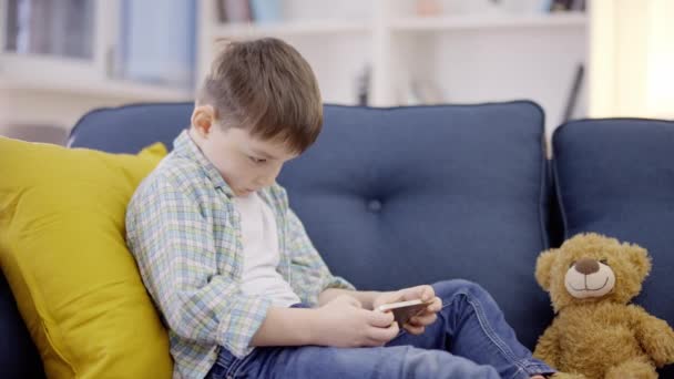 小男孩在智能手机上玩网络游戏 坐在家里的沙发上 小工具 — 图库视频影像
