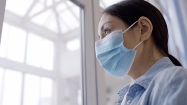 Hastane penceresinin yanında koruyucu maskeli düşünceli bir kadın duruyor.