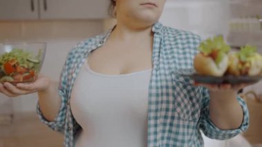 Şişman kadın, elinde salata, sosis ve kalori tutarak yemek seçimini düşünüyor.