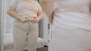 Şişko kadın, aynanın önünde dar pantolonunu kapatmaya çalışıyor.