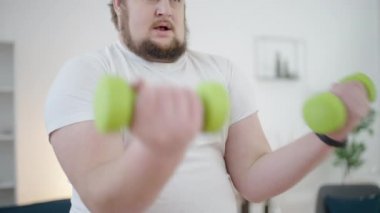 Yorgun obez adam evde çalışıyor, dambılları kaldırıyor, disiplin eğitimi alıyor.