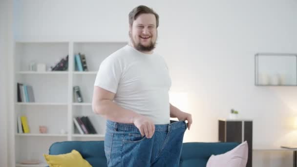 Šťastný mladý muž ukazuje nadměrné kalhoty, plán hubnutí, transformace