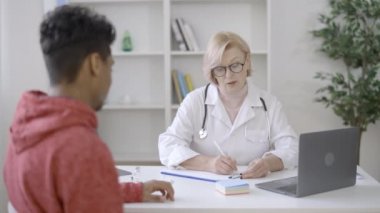 Genç erkek hasta klinikteki doktorla konuşuyor. Tıbbi kayıtları dolduruyor.