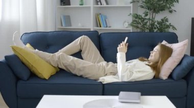 Gülümseyen genç kız kanepede dinleniyor, akıllı telefonda yazıyor, sosyal ağ uygulaması