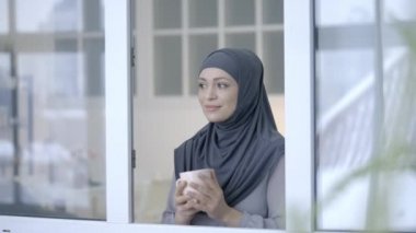 Güzel Müslüman kadın tesettürlü çay içiyor ve pencereden eve bakıyor.