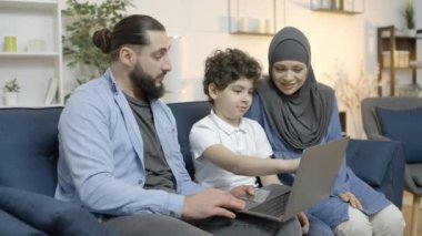 Neşeli Arap anne baba ve oğlu dizüstü bilgisayara bakıyor, birlikte tatil planlıyorlar.