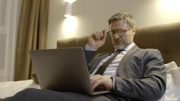 Konsultant Średnim Wieku Okularach Piszący Laptopie Siedzący Łóżku Nadgodziny — Wideo stockowe
