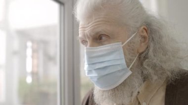 Koruyucu maskeli umutsuz yaşlı adam pencereden şehre bakıyor, Cavid-19.