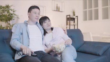 Genç Asyalı erkek ve kadın televizyon izliyor, abur cubur yiyor, birlikte dinleniyorlar.