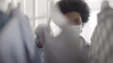 Maskeli ve eldivenli Afrikalı kadın Coronavirus sırasında kıyafetlere bakıyor, alışveriş yapıyor.