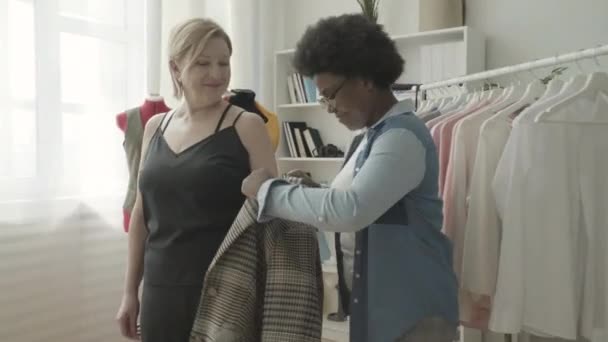 年轻的裁缝帮助顾客试穿夹克 快乐的顾客在时尚工作室里 — 图库视频影像
