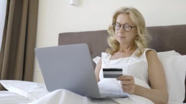 Memnun bir kadın kredi kartı bilgilerini bilgisayarda yatakta, internetten alışveriş yapıyor.