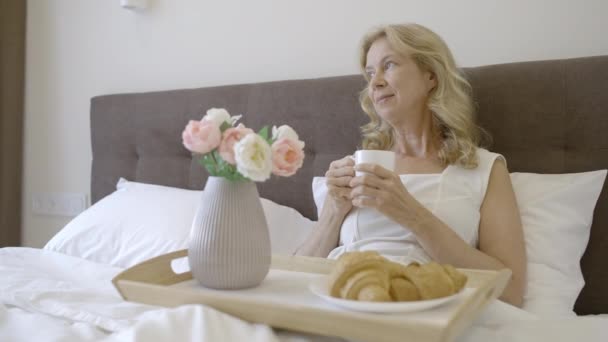 中年の女性がベッドで朝食をとり お茶を飲み 朝を楽しむ — ストック動画